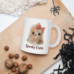 Op de afbeelding: Een witte keramische mok met een afbeelding van een geest gemaakt van patchworkstof met een roze strik en kanten rand. De tekst "Spooky Cutie" is in donkerrood onder de geest gedrukt. De mok heeft een standaard handvat en een gladde afwerking.