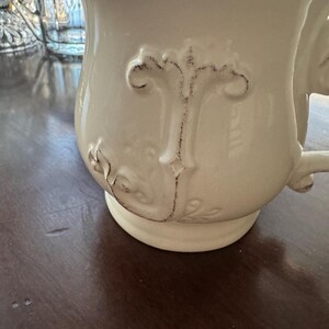 May include: Cream-colored ceramic mug with a decorative embossed design. The mug features a handle and a slightly flared rim with a distressed brown edge. The embossed design includes a fleur-de-lis and other floral elements.