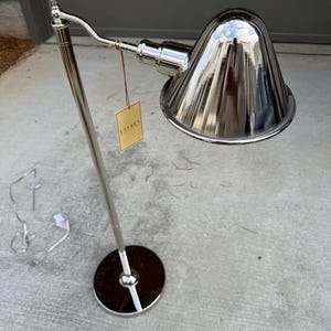 May include: A polished silver-tone metal floor lamp with a round base and a conical shade. The lamp features an adjustable arm and a rectangular tan tag hanging from the neck with the word LAUREN printed in black text. A thin power cord is attached.