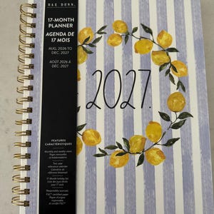 May include: A 17-month spiral-bound planner by Rae Dunn featuring a light blue and white vertical striped cover with a wreath of yellow lemons and green leaves. The cover displays the year 2027 and a black vertical band with product details.