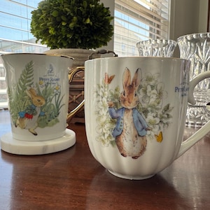 May include: Two white ceramic mugs with gold trim, featuring illustrations of Peter Rabbit. One mug has a tall, cylindrical shape, and the other is a large, rounded mug. Both mugs have the "Peter Rabbit" logo. The mugs are on a wooden table.