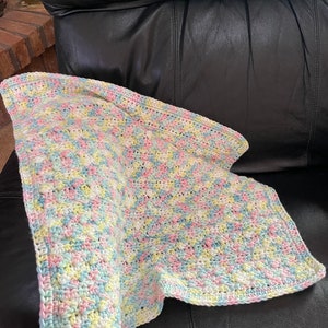 May include: A crocheted baby blanket in pastel shades of pink, blue, yellow, and white. The blanket is made with a textured stitch and has a soft, fluffy appearance.