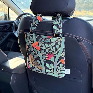 May include: A car seat organizer with a black background and a colorful botanical print featuring birds, berries, and flowers. The organizer hangs from the headrest and has a small tag on the bottom right corner.