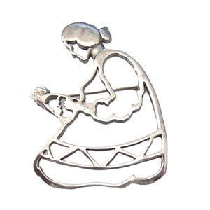May include: A silver-toned metal brooch in the shape of a woman kneeling and holding a baby. The brooch has an open design with cut-out details, and is set against a white background.
