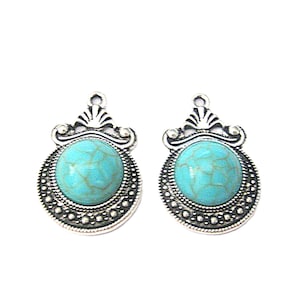 May include: Two silver-toned pendants with a turquoise-colored, round stone center. The pendants have a decorative border and a small loop at the top for attaching to a chain or other jewelry. The design is symmetrical and detailed.