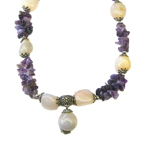 May include: A necklace featuring a combination of polished, light-colored stones and small, irregular purple amethyst chips. The necklace has a silver-colored clasp and a decorative pendant. The stones are strung together with silver-colored metal accents.