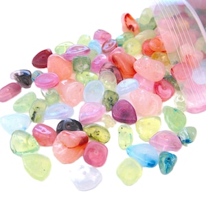 May include: A collection of small, tumbled, translucent beads in various colors including pink, green, blue, and yellow, spilling out from a clear plastic resealable bag. Each bead features a central hole, showcasing a smooth, polished surface.