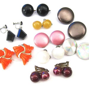 May include: A collection of vintage clip-on earrings in various styles and colors. Includes black faceted, yellow button, pink, white, iridescent, and burgundy pearl earrings. Also, orange fan-shaped and black geometric earrings.