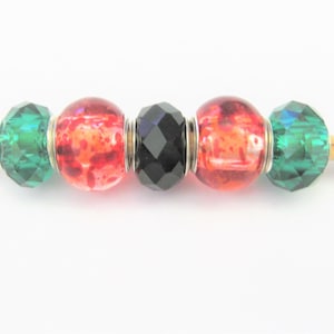 May include: A close-up of four beads on a wooden skewer. The beads are green, red with white speckles, black, and red with white speckles. The beads are all round and have a shiny finish.