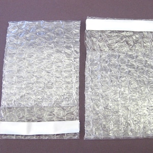 May include: Two clear bubble wrap pouches with white adhesive strips. The pouches are rectangular and filled with air bubbles. The pouches are designed for protecting items during shipping. The background is a solid, muted purple color.