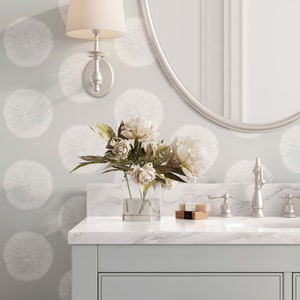 May include: A white bathroom vanity with a marble countertop, a silver faucet, and a glass vase with white flowers. The wall behind the vanity has a light gray wallpaper with a white floral pattern.