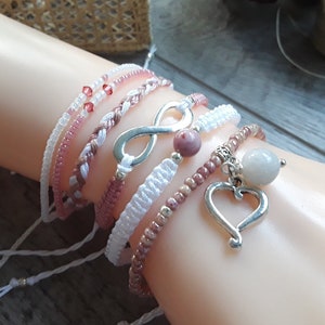 May include: A set of six bracelets with pink and white string, beads, and silver charms. The charms include an infinity symbol, a heart, and a round stone.