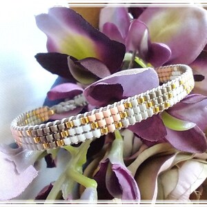 May include: A beaded bracelet with a light pink, white, and gray color scheme. The bracelet is made with small, round beads and has a simple, elegant design.