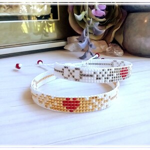 May include: Two white friendship bracelets with red and gold beads. The bracelets are woven with a heart design.