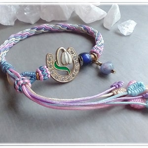 May include: A multi-colored braided bracelet with a silver charm featuring a white flower and green stem. The charm is surrounded by clear rhinestones. The bracelet has multiple tassels in pink, blue, and white.