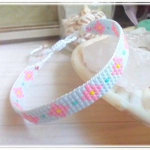 May include: A handmade friendship bracelet with a light blue background and a floral pattern in pink, yellow, and teal. The bracelet is adjustable with a white cord and features a small pink bead.