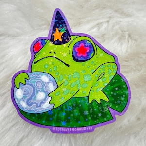 May include: A green frog wearing a purple and blue wizard hat with a yellow star on top, holding a blue and white orb. The frog is sitting on a purple lily pad with a green and blue glitter background. The text "#TOTALLYTHEGAMODPC" is at the bottom of the image.