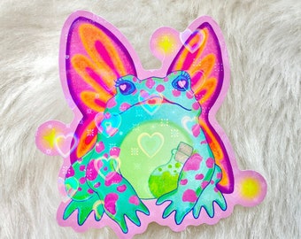 Puede incluir: Una pegatina con una rana verde con alas de mariposa rosa y naranja. La rana sostiene una botella de poción verde y está rodeada de corazones rosas.