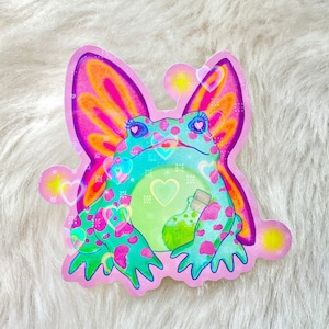 Op de afbeelding: Een sticker met een groene kikker met roze en oranje vlinder vleugels. De kikker houdt een groene drankfles vast en is omringd door roze harten.