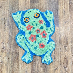 May include: A whimsical, tufted rug in the shape of a frog. The frog is primarily light green with blue accents and orange and blue eyes. Red flowers are scattered across the frog's body. The rug is placed on a wooden floor.