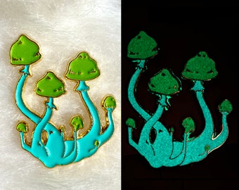 Puede incluir: Un pin de esmalte con forma de setas, con un borde dorado. Los tallos son de color azul claro y los sombreros de color verde brillante. El pin brilla en la oscuridad, mostrando un verde vibrante sobre un fondo negro. Un accesorio único.