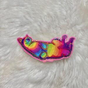 Pu&ograve; includere: Un adesivo olografico arcobaleno di un gatto che dorme sulla schiena. L'adesivo &egrave; su una superficie bianca e soffice.