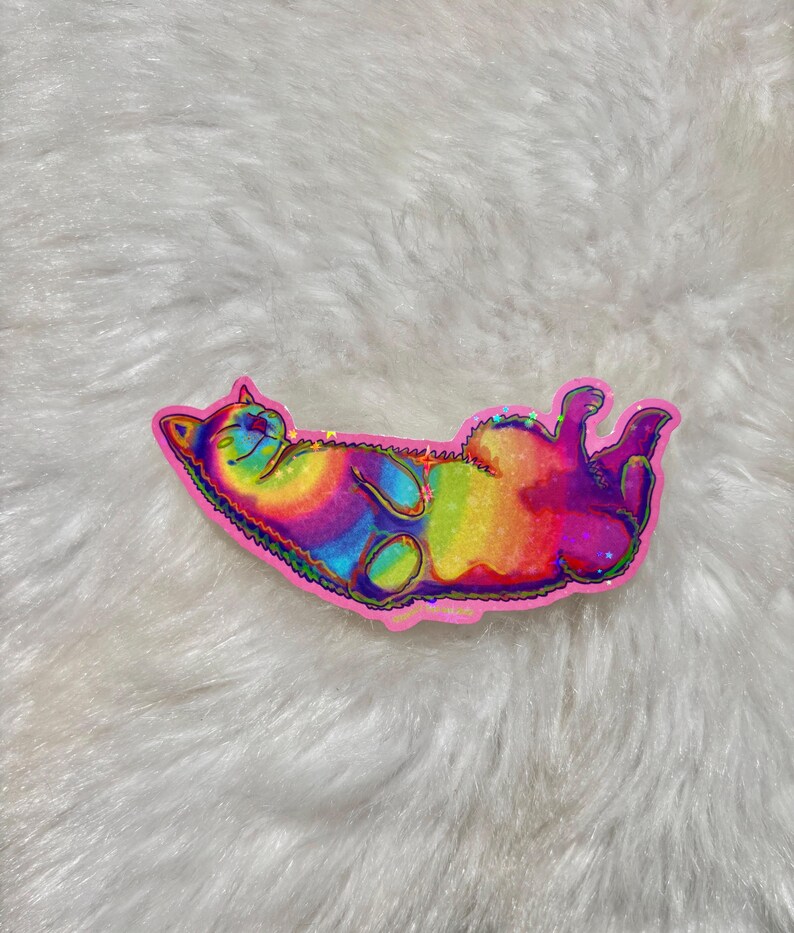 Pu&ograve; includere: Un adesivo olografico arcobaleno di un gatto che dorme sulla schiena. L'adesivo &egrave; su una superficie bianca e soffice.