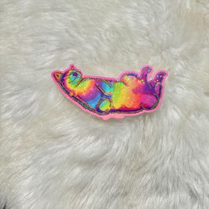 Pu&ograve; includere: Un adesivo olografico arcobaleno di un gatto che dorme sulla schiena. L'adesivo &egrave; su una superficie bianca e soffice.