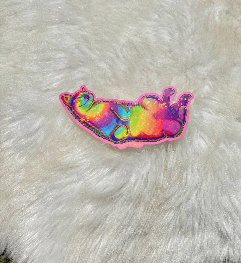 Pu&ograve; includere: Un adesivo olografico arcobaleno di un gatto che dorme sulla schiena. L'adesivo &egrave; su una superficie bianca e soffice.