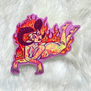 May include: A sticker featuring a woman with horns and a fiery halo. The woman is depicted in a reclining pose with her legs outstretched. The sticker has a holographic finish with a rainbow of colors.