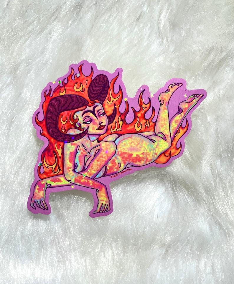 May include: A sticker featuring a woman with horns and a fiery halo. The woman is depicted in a reclining pose with her legs outstretched. The sticker has a holographic finish with a rainbow of colors.