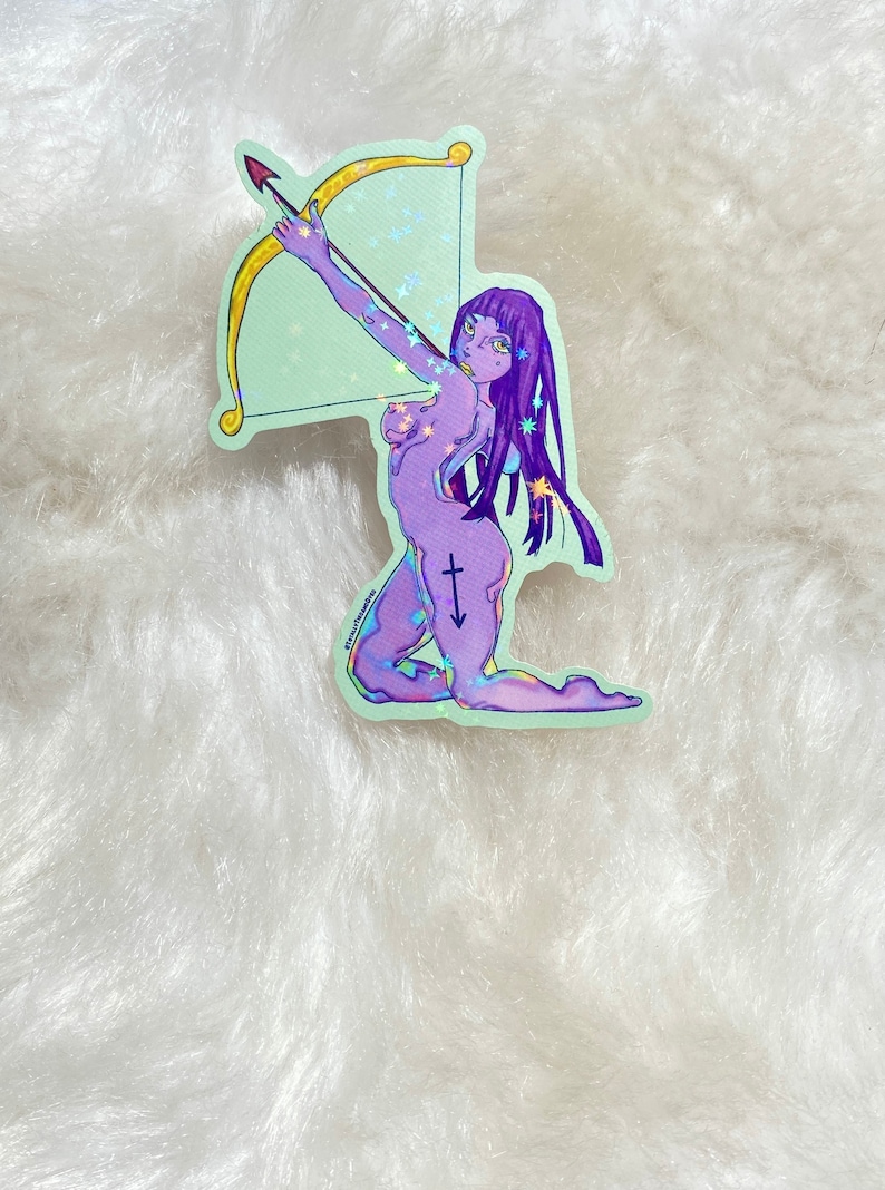 May include: A holographic sticker featuring a purple and blue illustration of a woman with long purple hair holding a bow and arrow. The woman is kneeling and has a Sagittarius symbol on her leg. The sticker has a light green background with glitter.