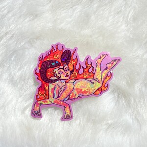 May include: A holographic sticker of a woman with horns and flames, lying on her side. The sticker is on a white fluffy surface. The sticker has a pink background and the text "@sweet.sundae" is in the bottom right corner.