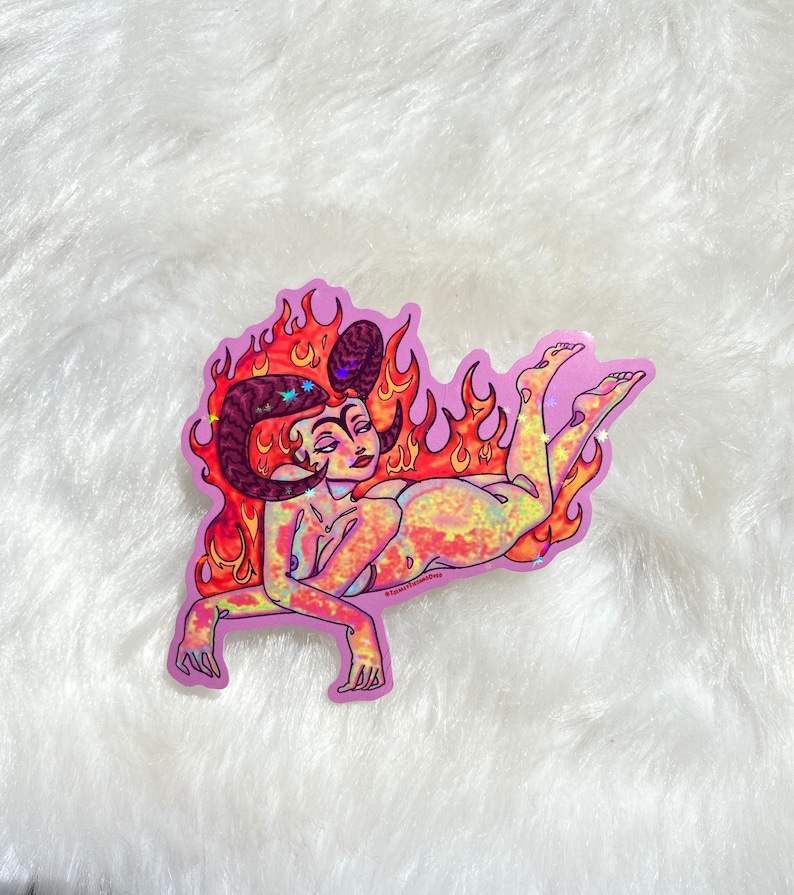May include: A holographic sticker of a woman with horns and flames, lying on her side. The sticker is on a white fluffy surface. The sticker has a pink background and the text "@sweet.sundae" is in the bottom right corner.