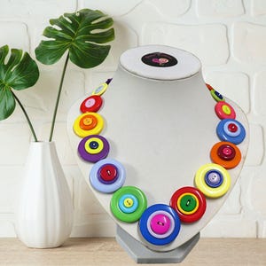 May include: A colorful button necklace displayed on a mannequin. The necklace features large, round buttons in various colors, including red, yellow, blue, green, and purple. Each button has a smaller button in the center. The necklace is a unique and artistic accessory.