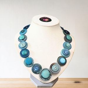 May include: A statement necklace featuring large, circular buttons in various shades of blue and teal. The buttons are arranged to create a bold design. The necklace is displayed on a white mannequin bust.