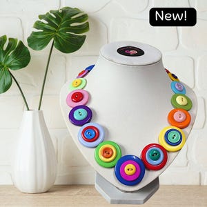 May include: A necklace featuring a series of layered, multi-coloured circular buttons in shades of red, blue, green, yellow, orange, and purple. The buttons are arranged in a graduated pattern on a white display bust with a black New! label.
