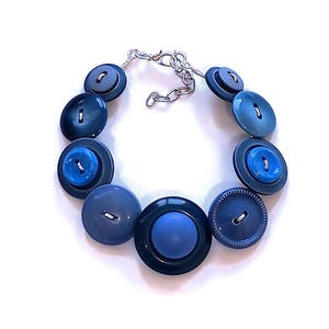 May include: A handcrafted bracelet composed of various blue buttons in diverse shades and dimensions. The buttons are linked by a silver-toned chain and clasp. This bracelet is a distinctive accessory.