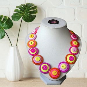 May include: A vibrant button necklace on a display bust. The necklace is made of layered buttons in pink, orange, yellow, and purple. The buttons are arranged in a circular design, creating a striking and playful accessory.