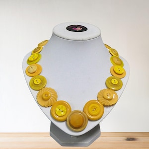 May include: A necklace composed of assorted yellow buttons, presented on a white mannequin bust. The buttons differ in size and tone, from light yellow to a richer, golden shade. This necklace is a distinctive, handmade jewellery item.