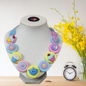 Könnte beinhalten: Ein farbenfrohes Collier auf einer Schaufensterpuppe. Die Halskette besteht aus großen, runden Knöpfen in Pastellfarben mit verschiedenen Designs, darunter Cupcakes und eine Teekanne. Die Knöpfe sind zu einem Schmuckstück verbunden.