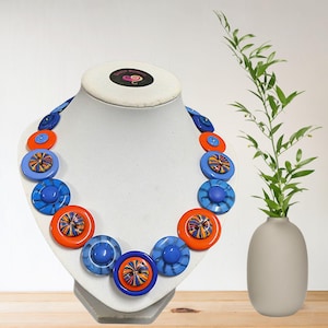 May include: A necklace made of large, colorful buttons. The buttons are blue, orange, and white with a variety of patterns. The necklace is displayed on a white mannequin bust.