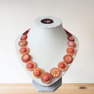 May include: A necklace composed of peach and pink buttons of varying sizes, displayed on a white mannequin bust. The buttons are arranged in a circular design. The necklace has a handcrafted appearance.