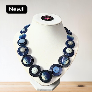 May include: A necklace composed of various circular buttons in shades of navy blue, light blue, and white, arranged in a graduated pattern. The buttons feature different textures, including smooth, ridged, and patterned surfaces, secured on a display.