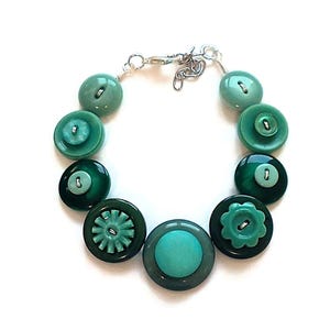 May include: A unique bracelet made from various green buttons, connected by a silver chain. The buttons vary in shade and size, creating a distinctive design. The bracelet's clasp is silver. The design showcases a mix of textures and shapes.