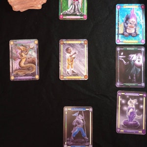 Pu&ograve; includere: Un set di nove carte da gioco fantasy con illustrazioni di varie creature e personaggi. Le carte sono bordate d'oro e hanno una finitura olografica. Le carte sono disposte in una griglia su una superficie nera.