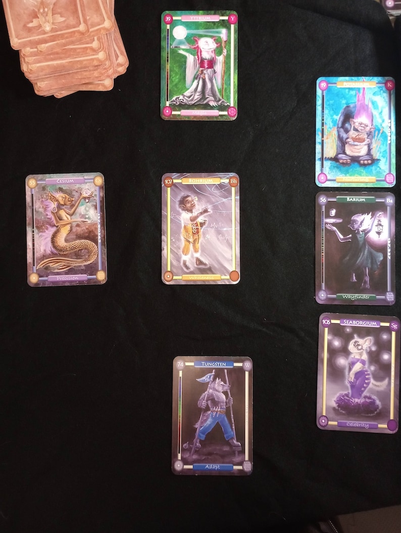 Pu&ograve; includere: Un set di nove carte da gioco fantasy con illustrazioni di varie creature e personaggi. Le carte sono bordate d'oro e hanno una finitura olografica. Le carte sono disposte in una griglia su una superficie nera.