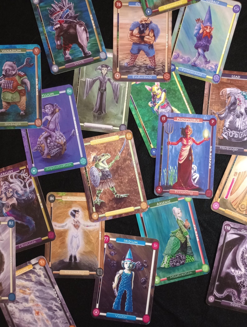 Pu&ograve; includere: Un set di carte da gioco colorate con creature fantastiche ed elementi. Ogni carta ha un'illustrazione e un testo unici, tra cui nomi come "Vanadium", "Polonium" e "Iridium".