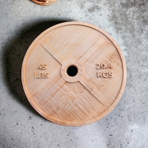 May include: A round, wooden weight plate with a central hole and engraved markings. The plate is light brown with visible wood grain. It features the numbers "45 LBS" and "20.4 KGS" carved into the surface, indicating weight measurements.