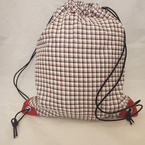 May include: A drawstring backpack with a white, black, and red plaid pattern. The bag has a red base and black drawstrings. The bag is closed at the top with a drawstring closure.